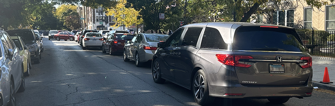 View of intersection with family posed to cross street, car traveling parallel to family, and cyclist riding on roadway.